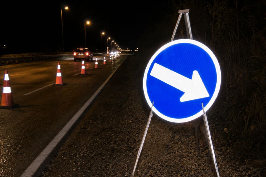 Señal azul de dirección obligatoria (flecha) instalada de noche en una carretera, con conos de tránsito y vehículos circulando
